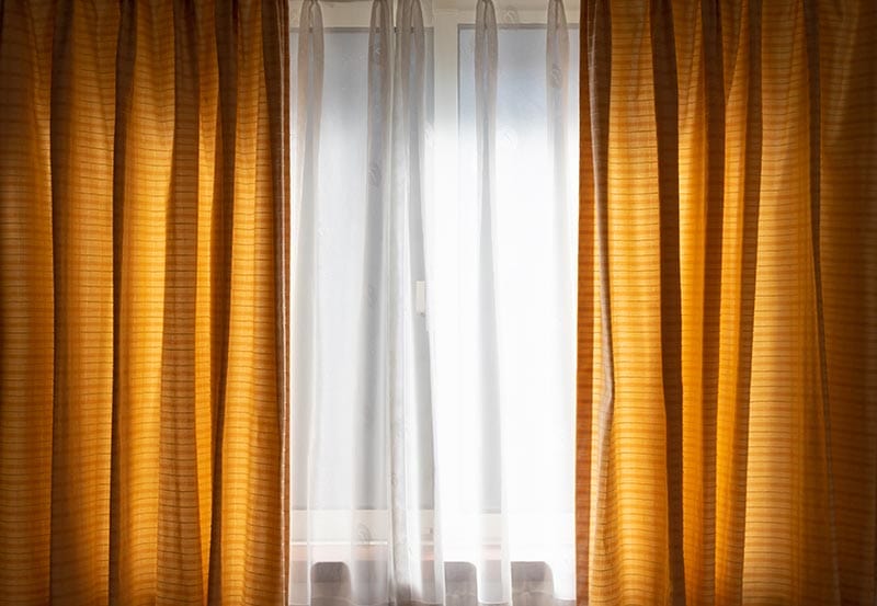 A bright window framed by heavy, golden-mustard textured curtains hanging on both sides, with sheer white drapes covering the center glass to soften the incoming daylight.