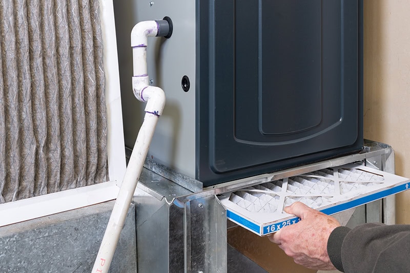 A hand sliding a clean, pleated air filter into the base slot of a home HVAC unit next to a dirty air filter.